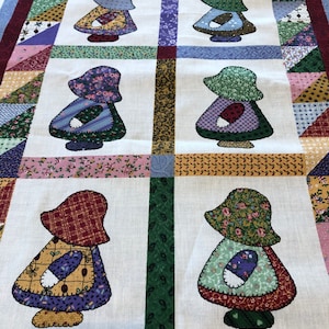 May include: A rectangular fabric quilt panel featuring six appliquéd figures wearing bonnets and dresses in various floral and geometric patterns. The central figures are arranged in two columns, surrounded by a border of colorful triangular fabric patches.