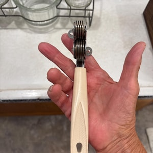 May include: A handheld kitchen tool featuring a cream-colored plastic handle with a textured leaf-like pattern and a hole at the base. The top consists of a metal sharpening mechanism with multiple stacked circular discs for blade maintenance.