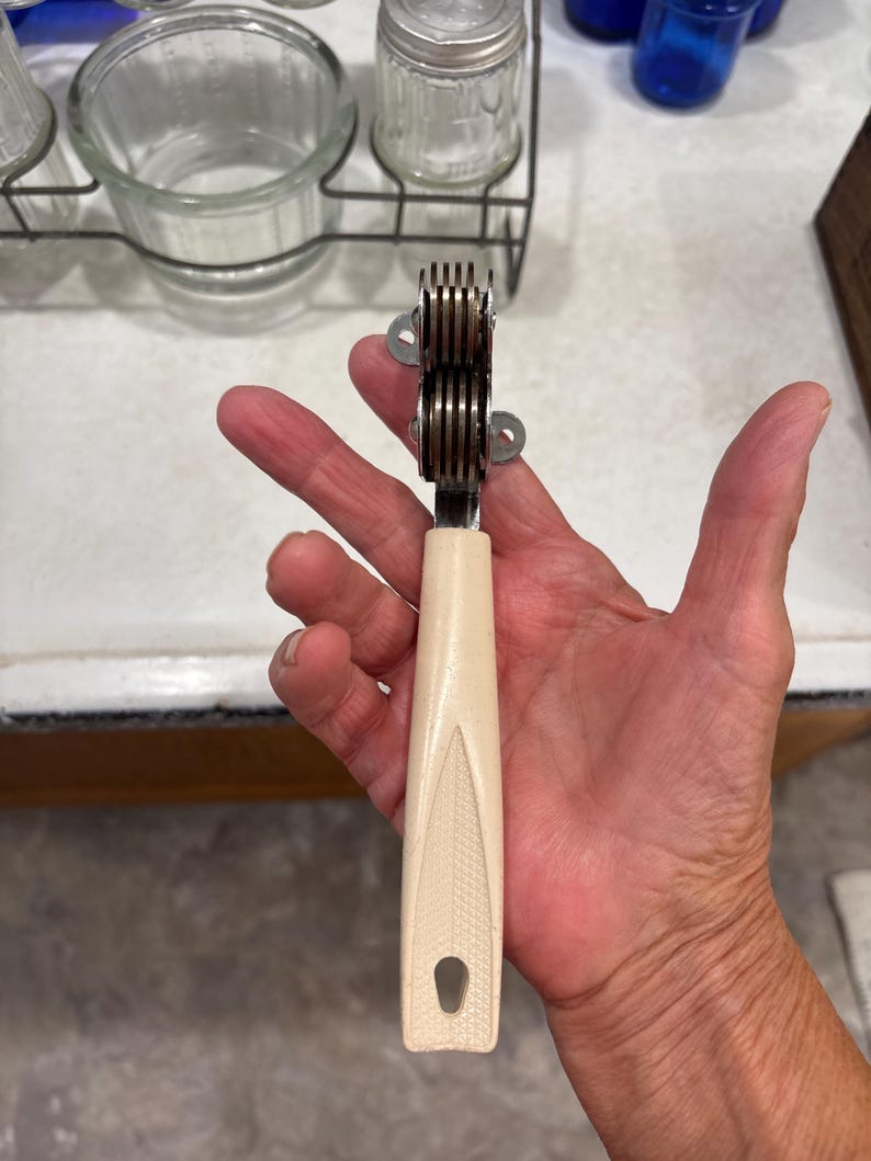 May include: A handheld kitchen tool featuring a cream-colored plastic handle with a textured leaf-like pattern and a hole at the base. The top consists of a metal sharpening mechanism with multiple stacked circular discs for blade maintenance.