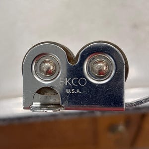 May include: A silver-colored metal kitchen tool featuring two circular wheels and a central housing stamped with the text EKCO U.S.A. The device has a smooth, reflective finish and a compact, functional design with visible rivets on each wheel.