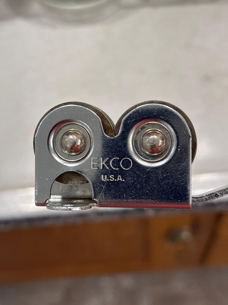 May include: A silver-colored metal kitchen tool featuring two circular wheels and a central housing stamped with the text EKCO U.S.A. The device has a smooth, reflective finish and a compact, functional design with visible rivets on each wheel.
