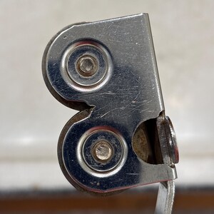 May include: A metal screen door roller assembly featuring two circular wheels secured within a silver-toned housing. The component includes a white plastic base attachment and shows visible signs of wear with surface scratches on the metal.