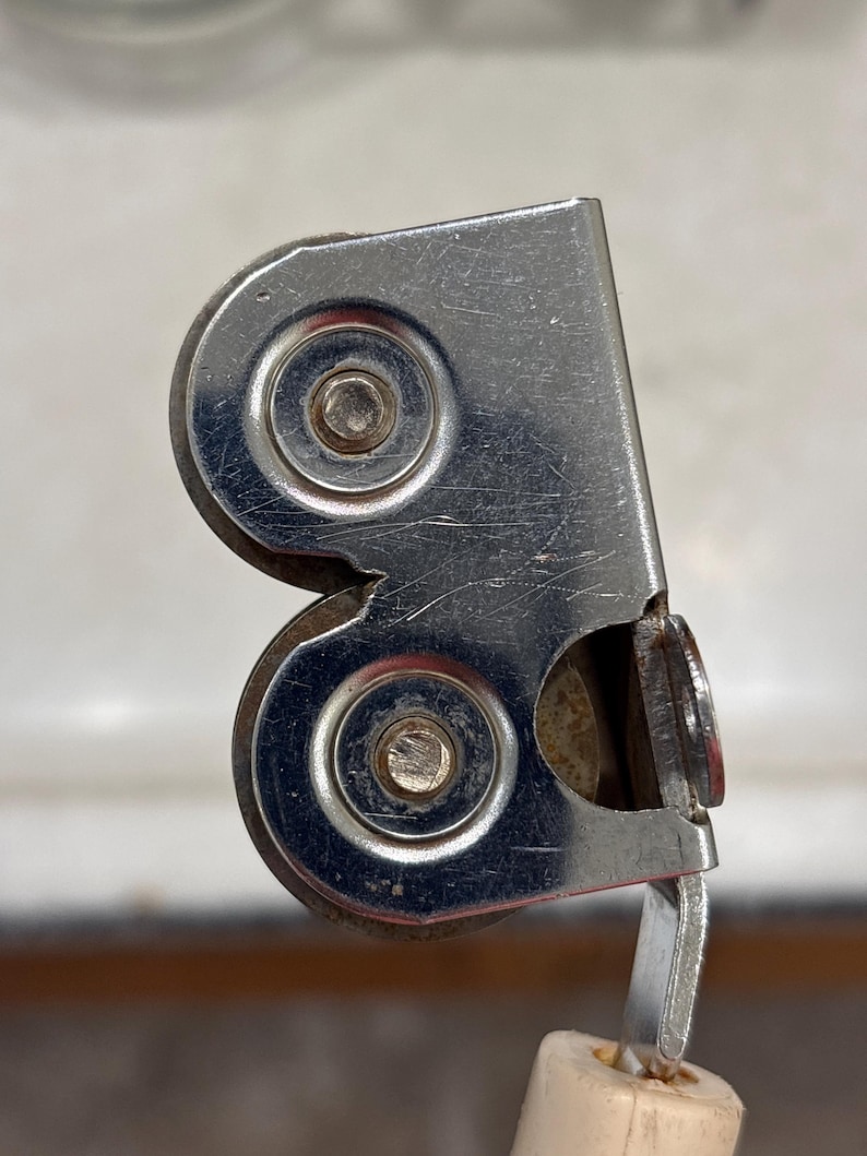 May include: A metal screen door roller assembly featuring two circular wheels secured within a silver-toned housing. The component includes a white plastic base attachment and shows visible signs of wear with surface scratches on the metal.