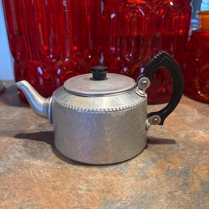 May include: A small, silver-colored metal teapot with a hammered texture on the body. It features a black plastic handle attached with metal screws, a matching black knob on the lid, and a beaded decorative band encircling the upper rim.