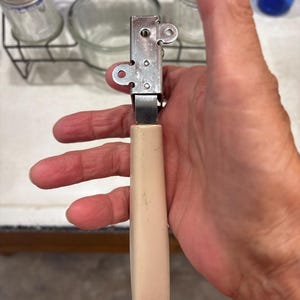 May include: A handheld kitchen tool featuring a rectangular stainless steel head with two circular side tabs and a central metal mechanism, attached to a long, smooth, off-white plastic handle with a small hole at the bottom end for hanging.