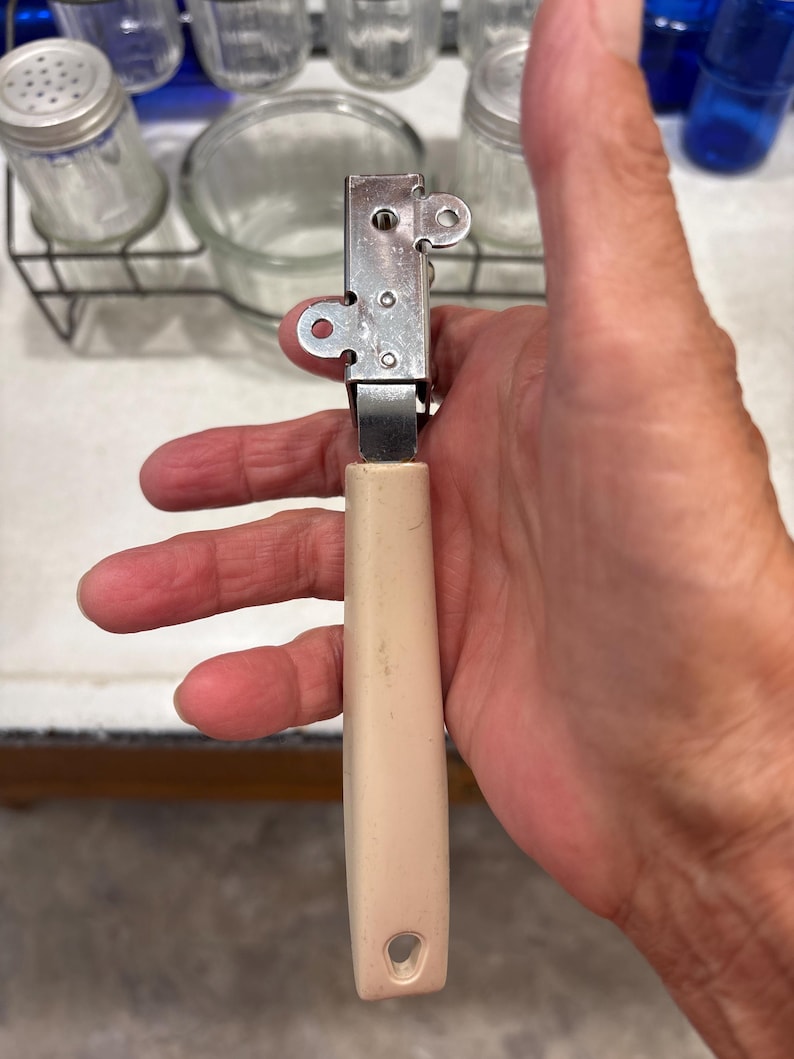 May include: A handheld kitchen tool featuring a rectangular stainless steel head with two circular side tabs and a central metal mechanism, attached to a long, smooth, off-white plastic handle with a small hole at the bottom end for hanging.