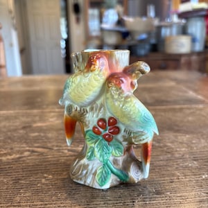 May include: A ceramic figurine of two parrots perched on a tree stump. The parrots have yellow, orange, and blue plumage. The tree stump is brown with green leaves and red berries. The figurine is on a wooden surface.