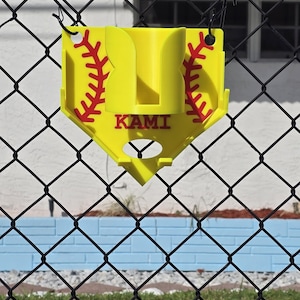 May include: A yellow softball holder with red stitching details, hanging on a chain-link fence. The holder has the word "KAMI" printed on it. The background includes a white building, blue wall, and green grass.