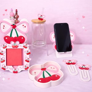 May include: A collection of cherry-themed desk accessories in pink and white. Includes a phone stand, memo holder, paper clips, and a small dish. A glass jar with a cherry straw and a pen holder are also present.