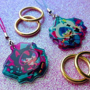 May include: An assortment of colorful anime-style charms and gold-colored rings. The charms feature vibrant illustrations with pink, teal, and black accents. The rings are circular and appear to be made of metal. The charms are attached to lanyards.
