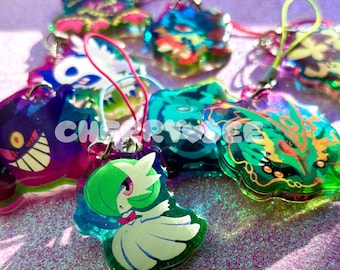 May include: A collection of acrylic charms featuring various colorful cartoon characters, including a green and white figure, a purple creature with a wide grin, and a green dragon-like form, each attached to a small colored loop for hanging.