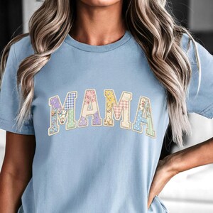 以下が含まれることがあります： フローラルとチェック柄のデザインで「MAMA」という文字が入った水色のTシャツ。文字はカレッジスタイルのフォントです。このシャツは半袖で、クルーネックです。