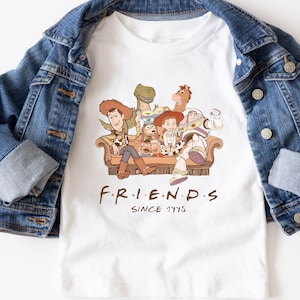 Pu&ograve; includere: T-shirt bianco con una grafica dei personaggi di Toy Story su un divano marrone, con la scritta "FRIENDS" e "SINCE 1995" stampata sotto. Una giacca di jeans blu &egrave; drappeggiata sulla maglietta.