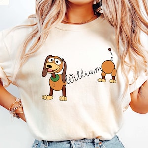 Puede incluir: Camiseta de color marfil con un diseño de perro de dibujos animados y el nombre "William" en fuente cursiva. El perro es marrón y canela con un collar verde. La camiseta es de estilo casual.