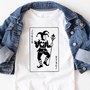 May include: White t-shirt featuring a black and white graphic of a joker playing card. The joker illustration includes a jester hat, heart, and scepter. A blue denim jacket is draped over the shirt.