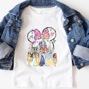 May include: White t-shirt featuring a colorful graphic of Disney princesses and a castle, framed by a Minnie Mouse bow. The shirt is paired with a blue denim jacket.