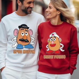 Puede incluir: Dos sudaderas, una blanca y otra roja, con diseños de personajes de dibujos animados de patatas. La blanca muestra "Spud Muffin", la roja "Sweet Potato". Los personajes llevan sombreros y bigotes.