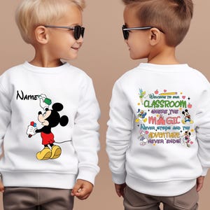 Puede incluir: Sudadera blanca con un dise&ntilde;o de Mickey Mouse sosteniendo un pincel y pintura. La parte delantera de la sudadera tiene la palabra "Name" encima del dise&ntilde;o de Mickey Mouse. La parte trasera de la sudadera tiene el texto "Welcome to our classroom where the magic never stops and the adventure never ends!"