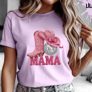 May include: Lilac t-shirt featuring a pink cowboy boot, hat, and disco ball design. The word "MAMA" is printed in pink, block letters. The shirt is a light purple color.
