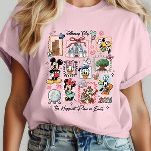 May include: A light pink t-shirt featuring a colorful graphic design with Disney characters and landmarks. The design includes Mickey Mouse, Minnie Mouse, Donald Duck, and Chip and Dale. Text on the shirt reads "Disney Trip" and "The Happiest Place on Earth" with the year "2026".