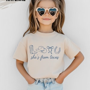 Puede incluir: Camiseta beige con un diseño azul de una bota de vaquero, un sombrero, un lazo y una herradura. El texto "she's from texas" está debajo. La palabra "NATURAL" está en la parte superior izquierda.