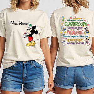 Puede incluir: Camiseta color crema con Mickey Mouse sosteniendo un rotulador rojo y el texto "Mrs. Name". La parte trasera de la camiseta tiene el texto "Welcome to our Classroom" con personajes de Disney y la frase "Never stops and the Adventure Never Ends!"