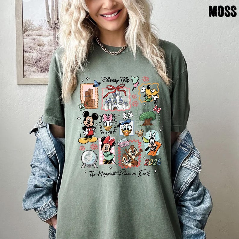 May include: Moss green t-shirt featuring a colorful Disney Trip graphic. The design includes various Disney characters, a castle, and the text "The Happiest Place on Earth" and "2026". The shirt has a relaxed fit.