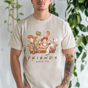 Pu&ograve; includere: T-shirt beige con un'illustrazione di personaggi di Toy Story seduti su un divano marrone. Il testo "FRIENDS SINCE 1995" &egrave; stampato sotto in marrone. Abbigliamento casual.