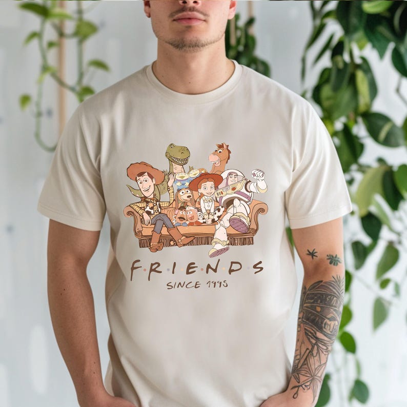 Pu&ograve; includere: T-shirt beige con un'illustrazione di personaggi di Toy Story seduti su un divano marrone. Il testo "FRIENDS SINCE 1995" &egrave; stampato sotto in marrone. Abbigliamento casual.