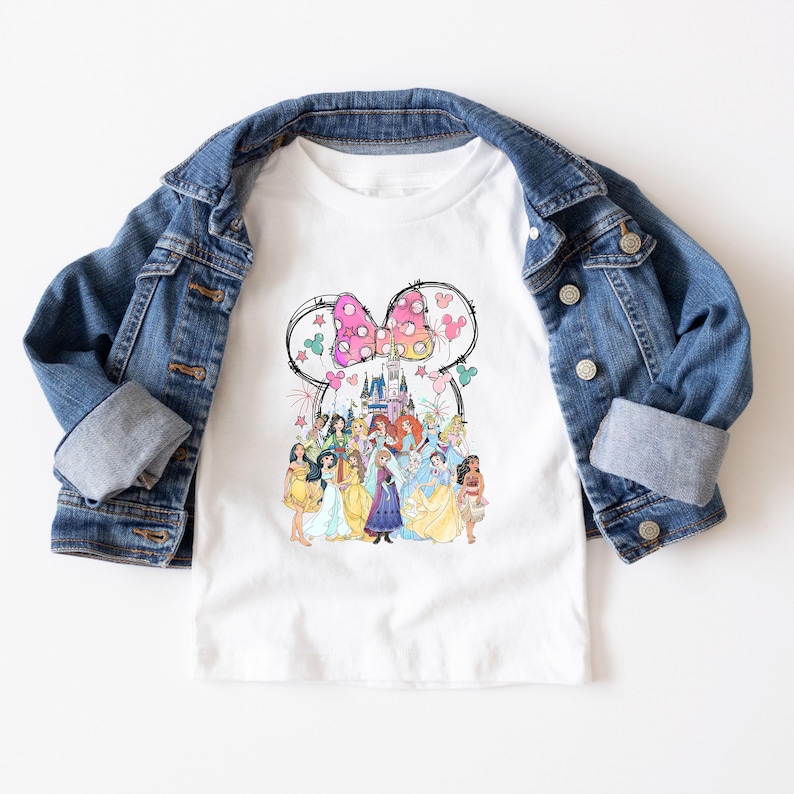 May include: White t-shirt featuring a colorful graphic of Disney princesses and a castle, framed by a Minnie Mouse bow. The shirt is paired with a blue denim jacket.