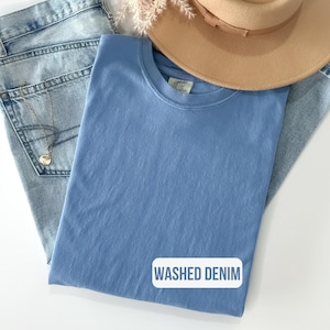 Puede incluir: Una camiseta de color azul b&iacute;garo liso con cuello redondo y etiqueta Comfort Colors en el interior del cuello. La prenda se coloca sobre unos vaqueros azul claro y un sombrero beige de ala ancha. Un recuadro blanco muestra WASHED DENIM.