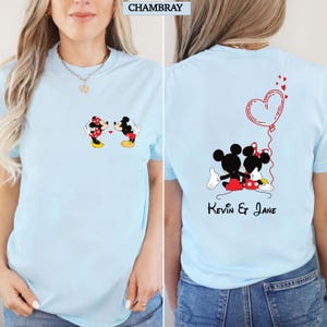 Puede incluir: Camiseta azul claro con ilustraciones de Mickey y Minnie Mouse. La parte delantera muestra a la pareja bes&aacute;ndose, mientras que la parte trasera los muestra abrazados con un globo en forma de coraz&oacute;n y los nombres "Kevin & Jane". La palabra "CHAMBRAY" est&aacute; arriba.