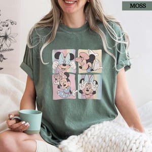 Puede incluir: Camiseta verde musgo con un gr&aacute;fico de cuatro paneles de Minnie Mouse en varias poses. Cada panel tiene un fondo pastel. La camiseta tiene mangas cortas y un corte holgado. La palabra "MOSS" est&aacute; impresa en un recuadro en la esquina superior derecha.