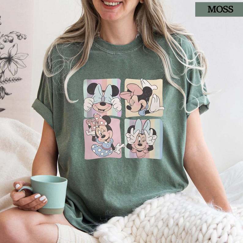Puede incluir: Camiseta verde musgo con un gr&aacute;fico de cuatro paneles de Minnie Mouse en varias poses. Cada panel tiene un fondo pastel. La camiseta tiene mangas cortas y un corte holgado. La palabra "MOSS" est&aacute; impresa en un recuadro en la esquina superior derecha.