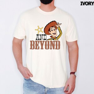 May include: Ivory t-shirt with a cartoon cowboy graphic, brown hat, and the text "AND BEYOND". The cowboy is waving, and a gold sheriff's star is next to the text. The shirt is a light cream color.