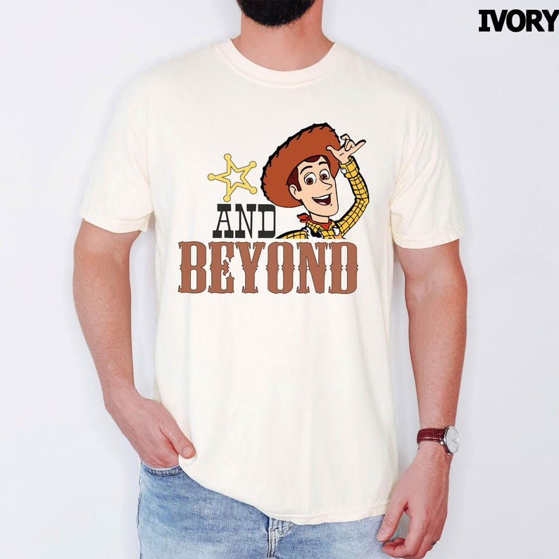 May include: Ivory t-shirt with a cartoon cowboy graphic, brown hat, and the text "AND BEYOND". The cowboy is waving, and a gold sheriff's star is next to the text. The shirt is a light cream color.