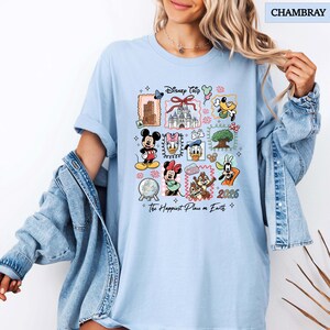 May include: Light blue t-shirt with a colorful Disney Trip graphic. The design features various Disney characters and landmarks, including Mickey Mouse, Minnie Mouse, and Cinderella's castle. The text "2026" and "The Happiest Place on Earth" are also included.