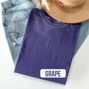 Puede incluir: Una camiseta de cuello redondo de color uva s&oacute;lido con la palabra GRAPE impresa en una etiqueta rectangular blanca en la parte inferior. La camiseta est&aacute; sobre unos vaqueros azul claro y parcialmente cubierta por un sombrero beige de ala ancha con banda marr&oacute;n.