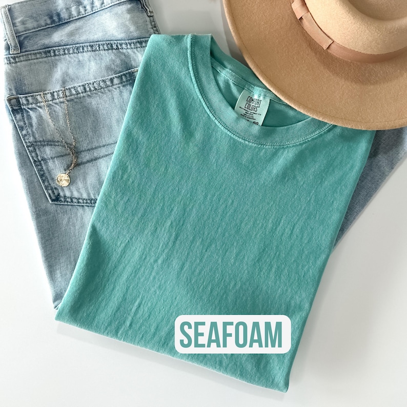Puede incluir: Una camiseta de cuello redondo color verde espuma de mar con etiqueta Comfort Colors, combinada con jeans azul claro, un sombrero beige de ala ancha y un collar de cadena dorada. El texto SEAFOAM est&aacute; impreso en blanco sobre un fondo rectangular.