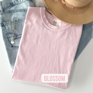 Puede incluir: Una camiseta rosa claro con una etiqueta de la marca Comfort Colors en el cuello y la palabra BLOSSOM impresa en rosa sobre un fondo rectangular blanco. La camiseta se superpone a unos vaqueros azul claro, un sombrero beige y un collar.