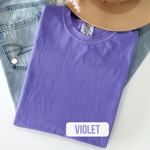 Puede incluir: Una camiseta de color violeta s&oacute;lido de manga corta con cuello redondo. La etiqueta interior del cuello muestra la marca Comfort Colors y la talla XL-TG-EG. La tela tiene una apariencia ligeramente texturizada y te&ntilde;ida con un tono p&uacute;rpura uniforme.