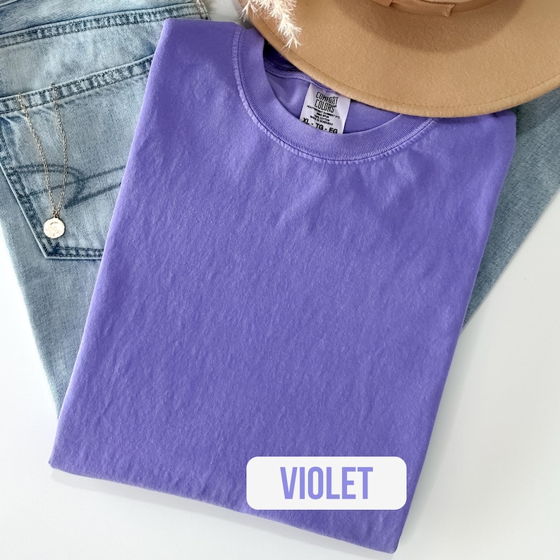 Puede incluir: Una camiseta de color violeta s&oacute;lido de manga corta con cuello redondo. La etiqueta interior del cuello muestra la marca Comfort Colors y la talla XL-TG-EG. La tela tiene una apariencia ligeramente texturizada y te&ntilde;ida con un tono p&uacute;rpura uniforme.