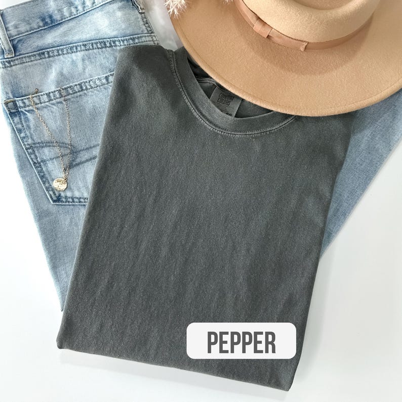 Puede incluir: Una camiseta de cuello redondo gris carb&oacute;n con etiqueta Comfort Colors, vaqueros azul claro, un sombrero beige de ala ancha y un collar de cadena dorada con un peque&ntilde;o colgante circular. Se ve una etiqueta blanca con el texto PEPPER.