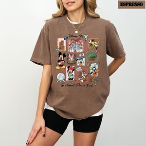 May include: Espresso-colored t-shirt featuring a colorful Disney Trip design. The design includes various Disney characters and landmarks, with the text "Disney Trip" and "The Happiest Place on Earth". The year "2026" is also visible.