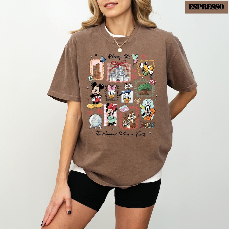 May include: Espresso-colored t-shirt featuring a colorful Disney Trip design. The design includes various Disney characters and landmarks, with the text "Disney Trip" and "The Happiest Place on Earth". The year "2026" is also visible.