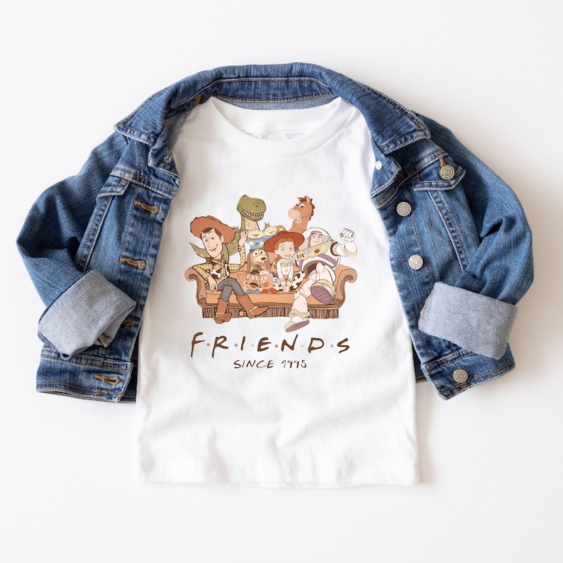 Pu&ograve; includere: T-shirt bianco con una grafica dei personaggi di Toy Story su un divano marrone, con la scritta "FRIENDS" e "SINCE 1995" stampata sotto. Una giacca di jeans blu &egrave; drappeggiata sulla maglietta.