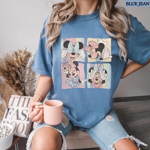 Puede incluir: Camiseta azul con un gr&aacute;fico de cuatro paneles de Minnie Mouse en varias poses. Cada panel tiene un fondo de arco&iacute;ris pastel. La camiseta tiene mangas cortas y un corte holgado. El texto "BLUE JEAN" est&aacute; en la esquina superior derecha.