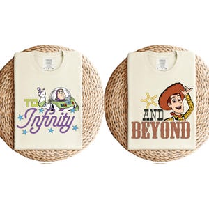 May include: Two cream-coloured t-shirts with cartoon graphics. One features Buzz Lightyear with the text "To Infinity" in purple script. The other shirt displays Woody with the text "And Beyond" in brown letters.