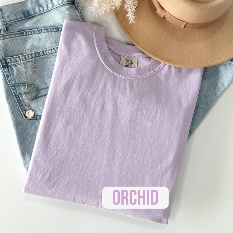 Puede incluir: Una camiseta de cuello redondo en color orqu&iacute;dea s&oacute;lido con una etiqueta de la marca Comfort Colors en el interior del cuello. La prenda se muestra junto a unos vaqueros azul claro, un sombrero beige de ala ancha y un collar de cadena plateada.