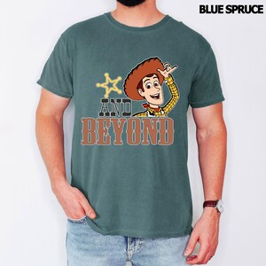 May include: Blue Spruce t-shirt with a cartoon image of Woody from Toy Story. The design includes a sheriff's badge, the words "AND BEYOND" in brown, and Woody. The shirt is a solid colour and has short sleeves.